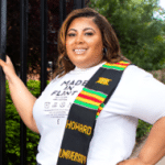 smiling woman with brown hair wearing a white shirt and colorful sash standing outside