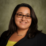 profile photo of smiling woman with long dark hair, dark-rimmed glasses, a black suit jacket and yellow shirt against a brownish background